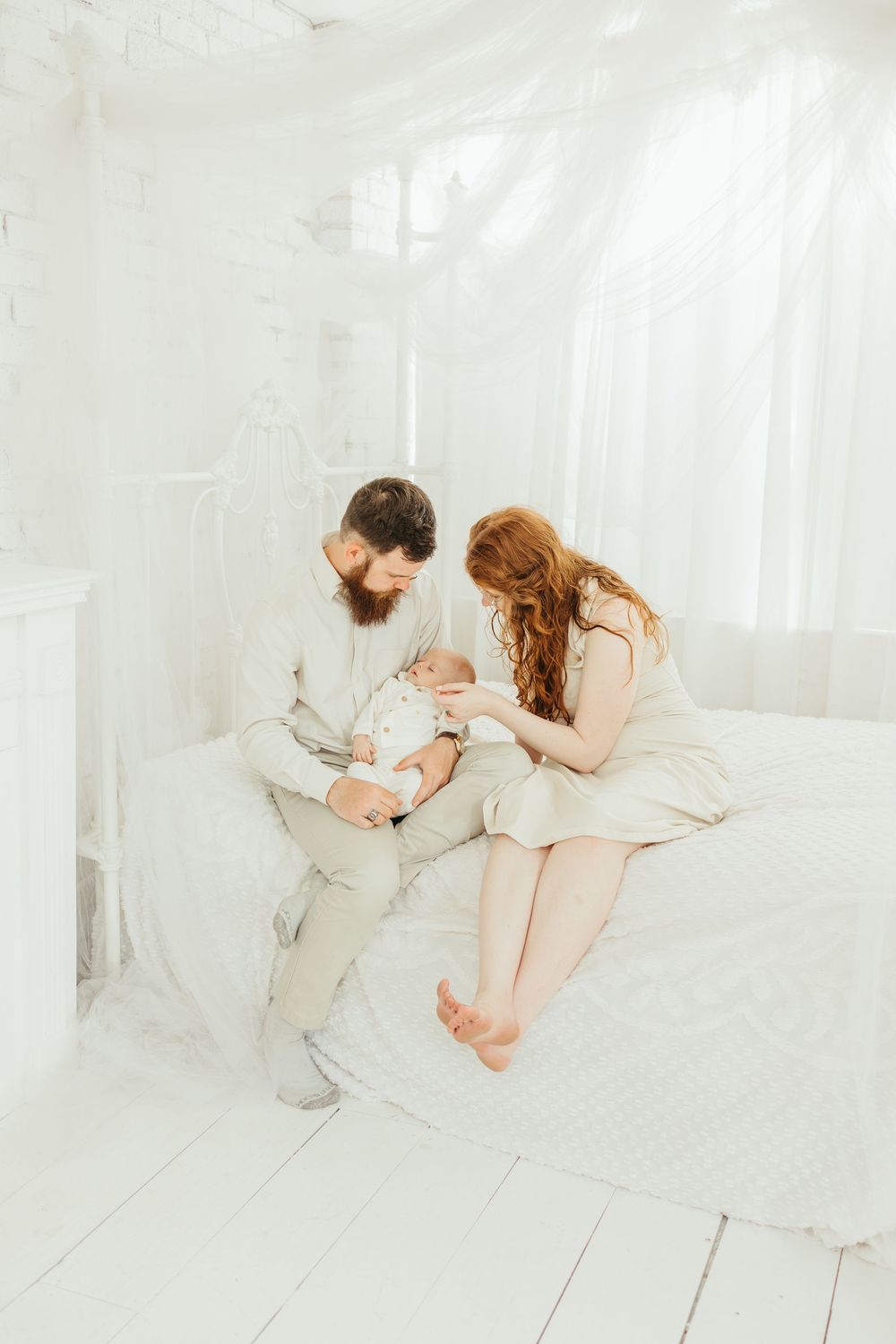 A bearded man and redhead woman sit on a white bed together, lovingly cradling their newborn baby in a bright room.
