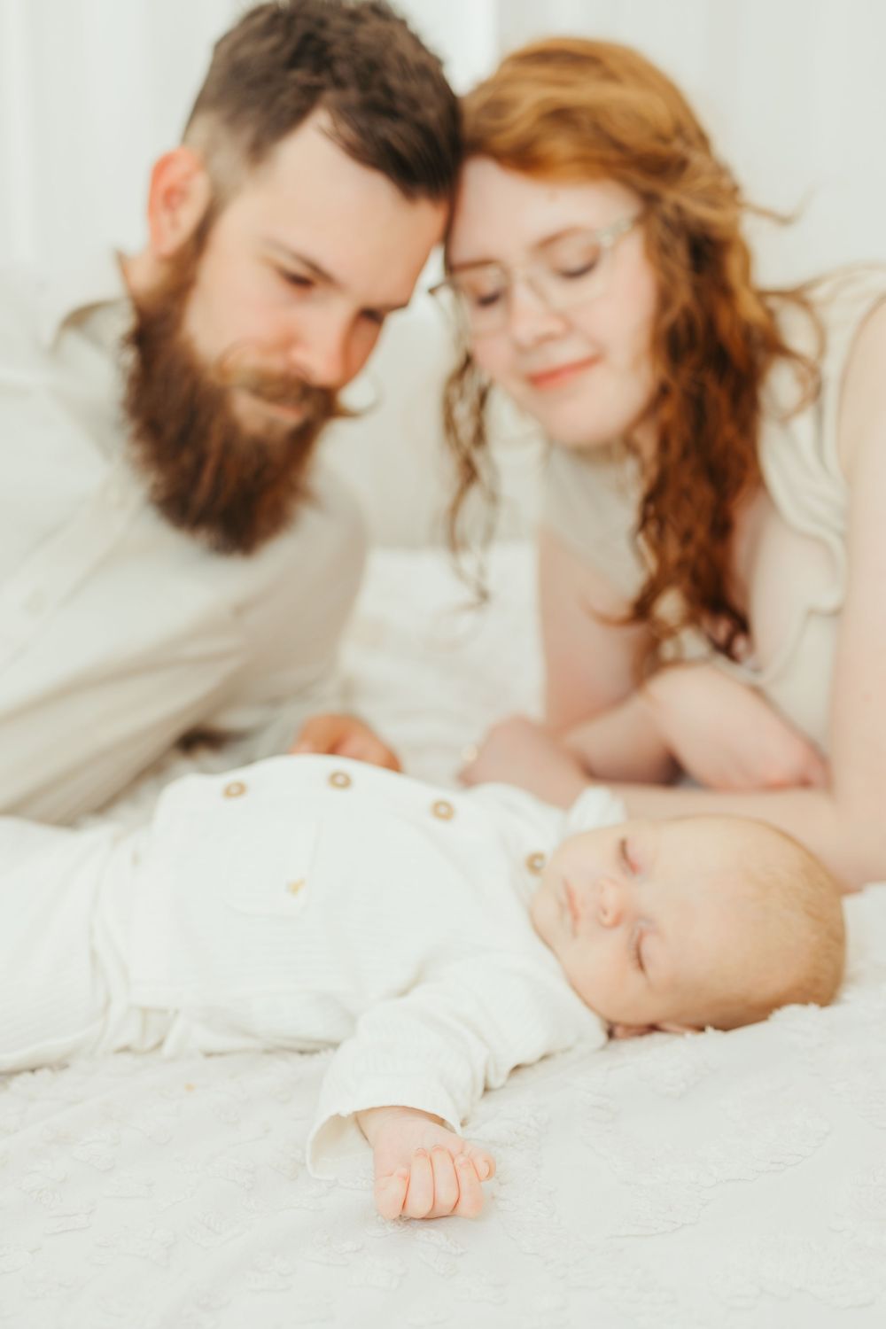 Bearded father and redhead mother lovingly gaze at their sleeping newborn baby dressed in white on a soft bed.