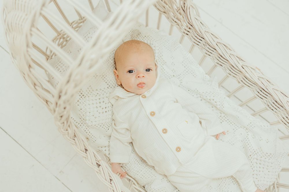 Newborn baby in white knit outfit lying in a wicker bassinet on a white background.