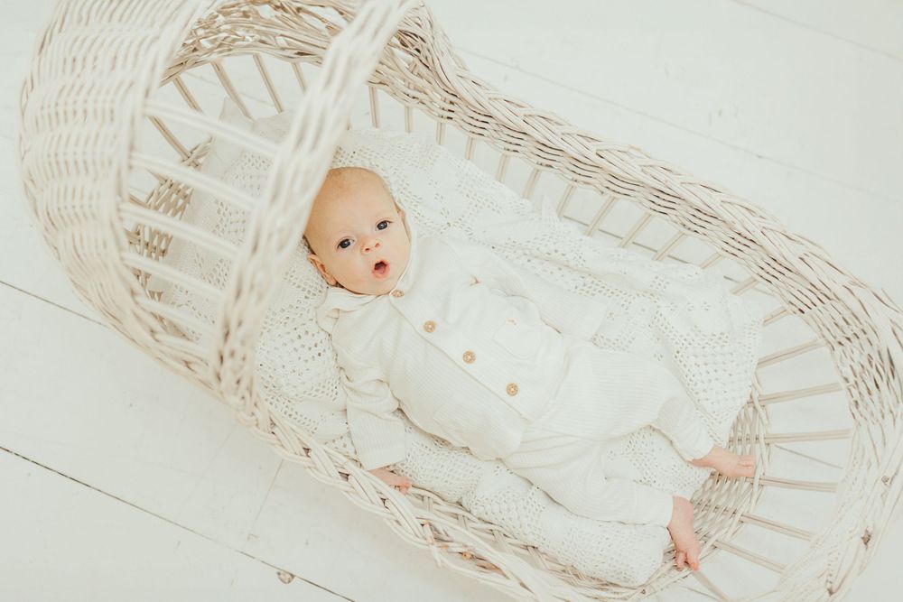 A peaceful newborn baby in a white knit outfit lying in a wicker Moses basket on a white floor.