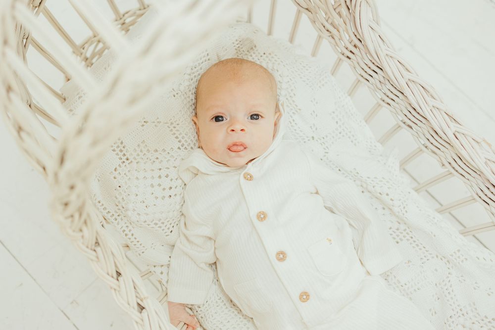 A sweet baby in a white knit outfit and bonnet lying in a wicker bassinet, gazing upward with bright eyes.