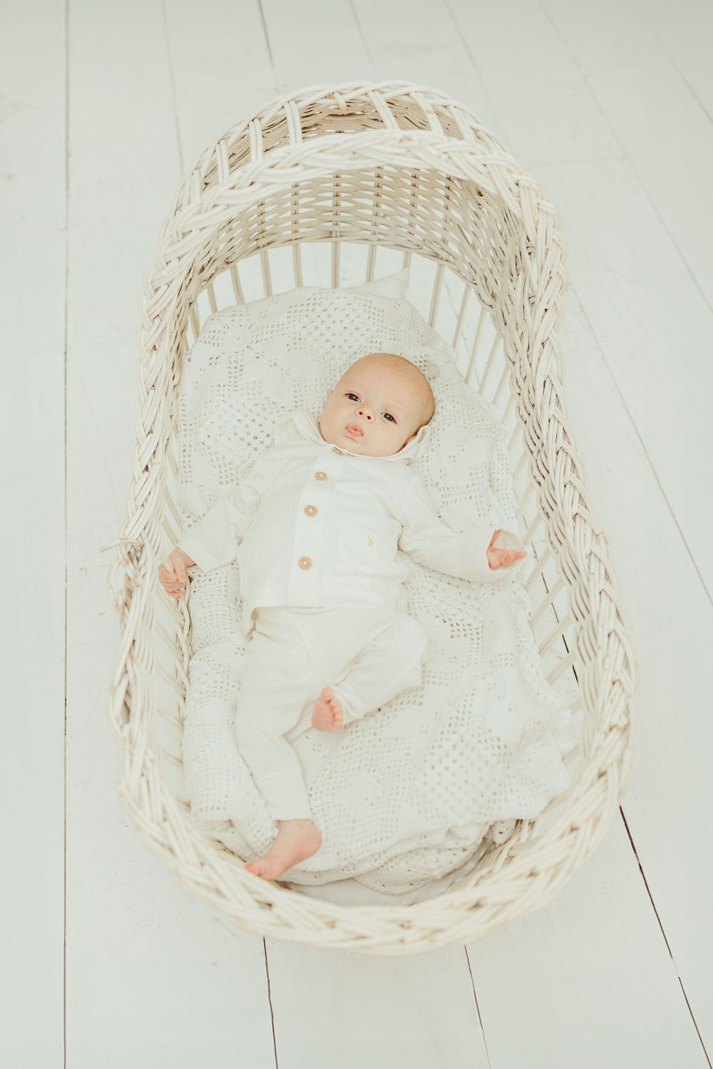 A peaceful baby in white outfit lying in a wicker bassinet on a white knit blanket.