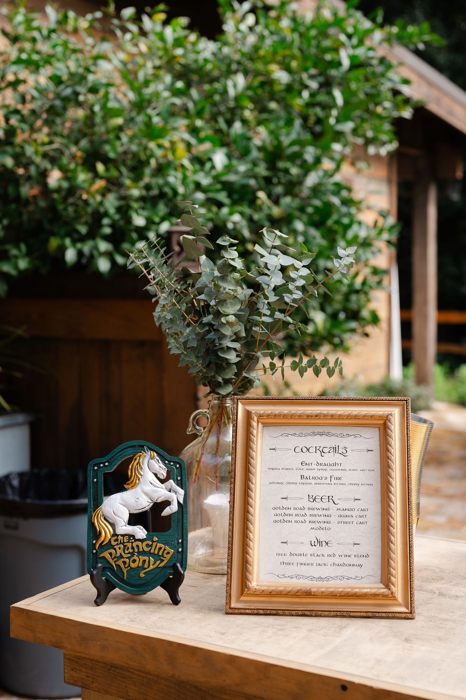 Lord of the Rings themed wedding bar menu in gold frame beside The Prancing Pony sign with eucalyptus centerpiece.
