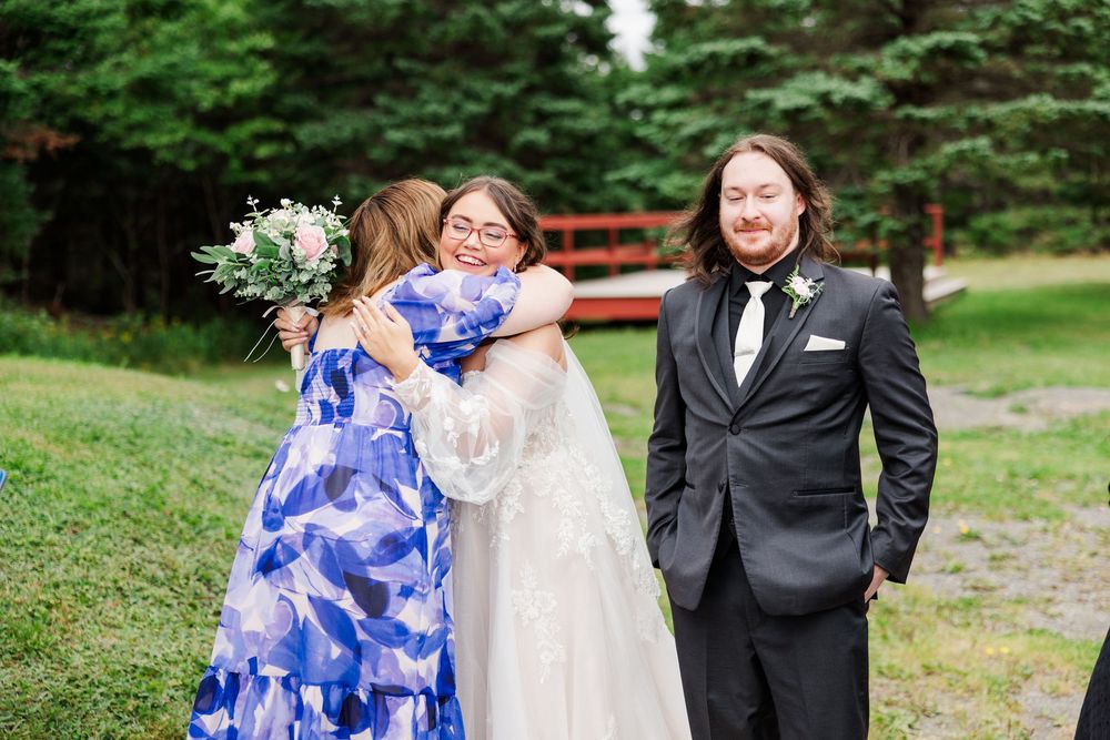 Bride in white gown hugging guests in blue floral dress while groom in black suit smiles at outdoor wedding.