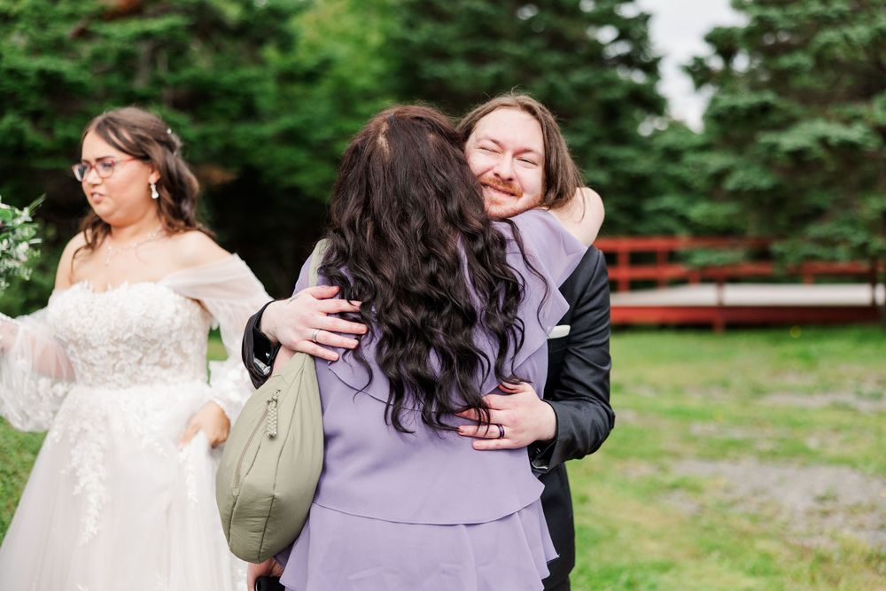 Two women share a warm hug at a wedding while a bride in a white gown stands nearby outdoors.