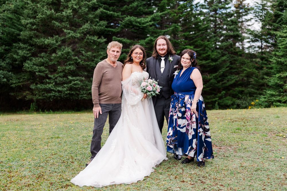 Wedding couple in white gown and suit pose with two guests outdoors near evergreen trees on a grassy lawn.
