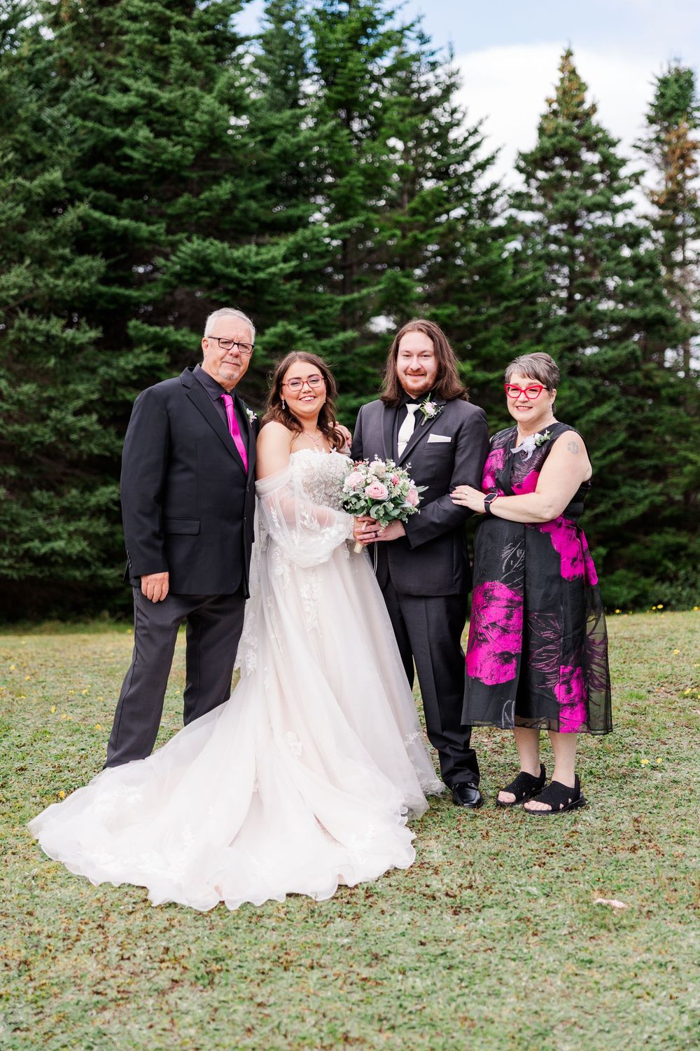 Wedding party of four posing outdoors, bride in white gown with bouquet, groom in black tux, guests in pink accents.