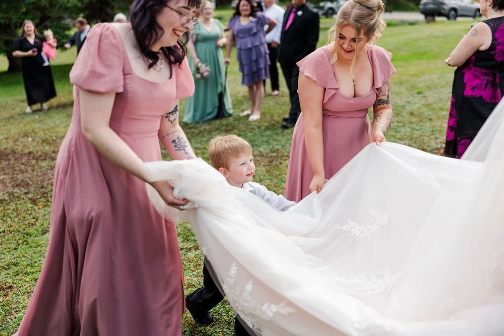 Bridesmaids in pink dresses help a young boy with a white wedding dress train at an outdoor wedding.