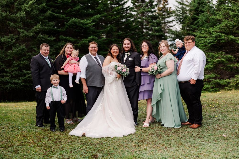 Wedding party poses outdoors on green lawn with evergreen trees, bride in white gown surrounded by family in formal attire.