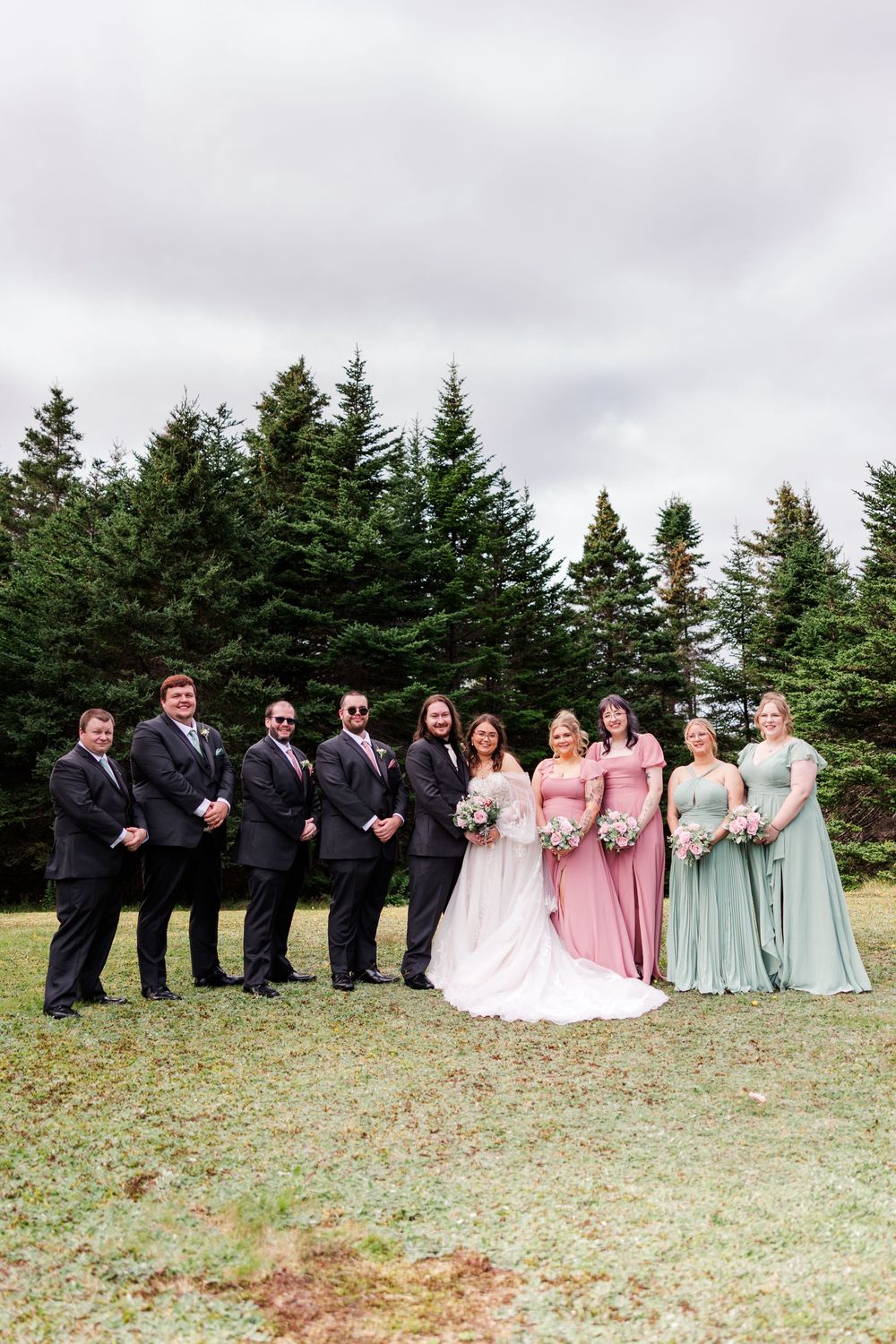 Wedding party poses outdoors with evergreen trees, groomsmen in black suits and bridesmaids in pink and sage green gowns.