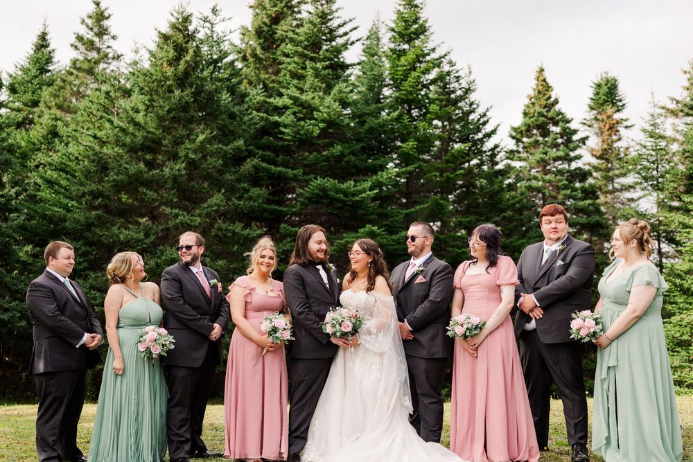 Wedding party posing outdoors with evergreen trees, bride in white gown flanked by pink and sage green bridesmaids and groomsmen.