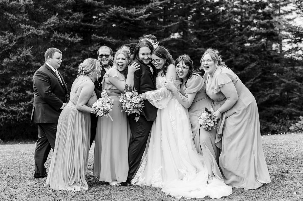 Black and white wedding photo of bride and groom surrounded by laughing bridesmaids and groomsmen outdoors.