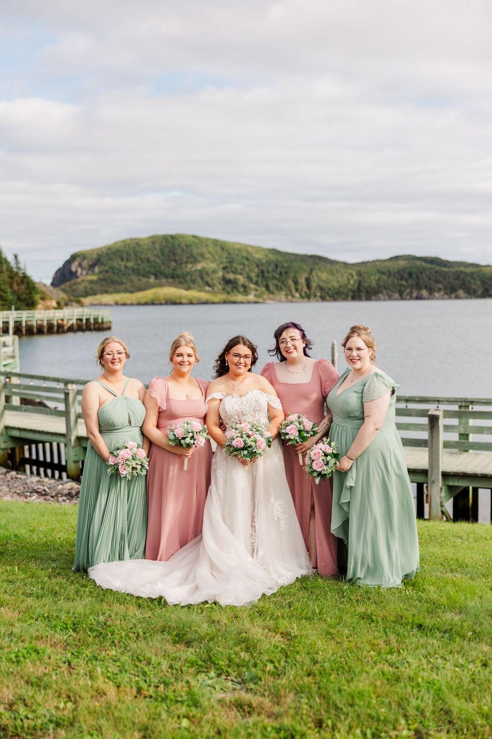 Bride in white gown poses with four bridesmaids in sage green and dusty rose dresses, holding bouquets by a scenic waterfront.