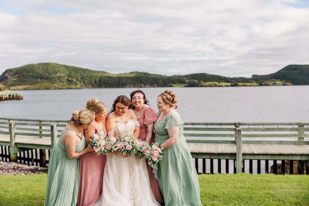 Bride and bridesmaids in sage green and dusty rose gowns laughing together outdoors by a scenic lake and green hills.