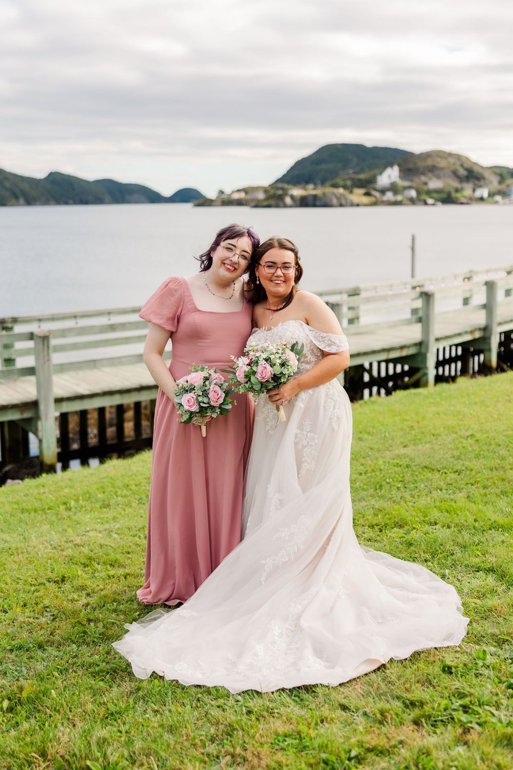 Bride in white gown and bridesmaid in pink dress holding bouquets by waterfront with mountains backdrop.
