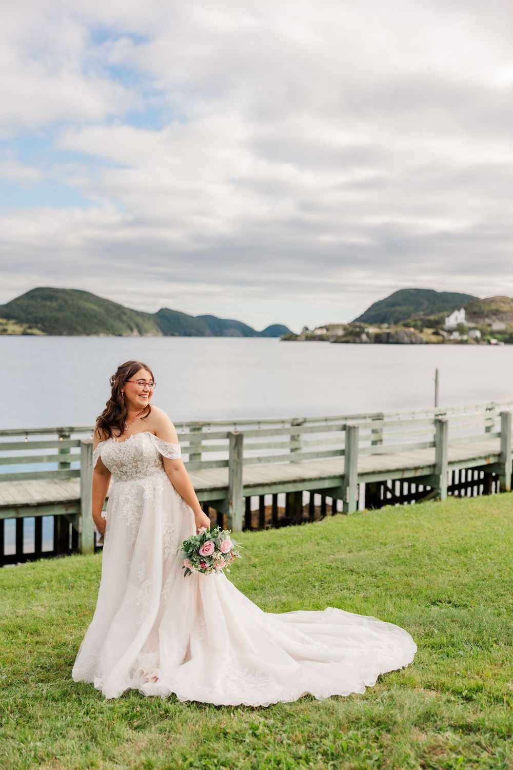 Smiling bride in off-shoulder lace gown holds bouquet by waterfront dock with misty hills backdrop.