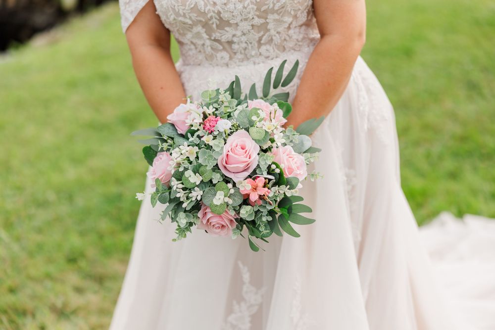 Bride in lace wedding gown holding a pink rose and greenery bridal bouquet outdoors on green grass.