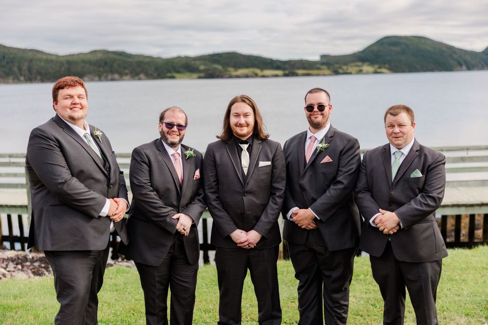 Five groomsmen in dark suits with pink ties pose outdoors near a scenic lake and green hills on a wedding day.