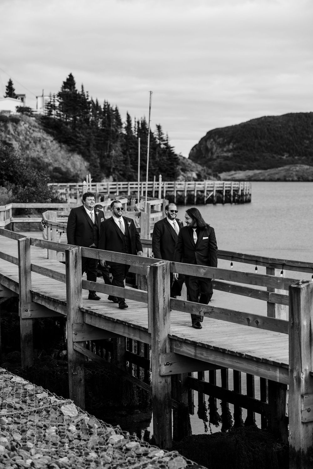 Black and white photo of groomsmen walking on a wooden dock by the water, trees and hills in the background.