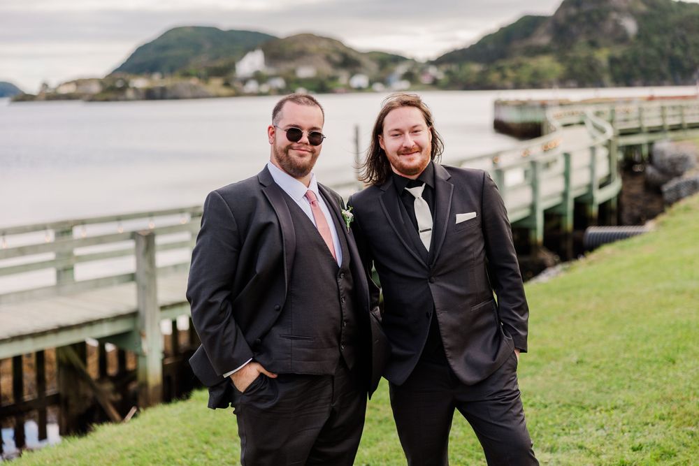 Two grooms in black suits pose smiling outdoors near a waterfront dock with green hills in the background.