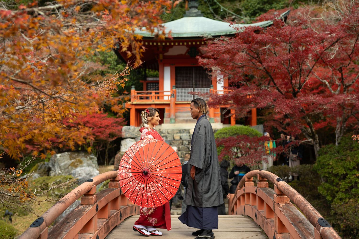 Man kneeling on a red wooden bridge proposing to a woman in a white dress, with a traditional Japanese temple and autumn foliage behind.