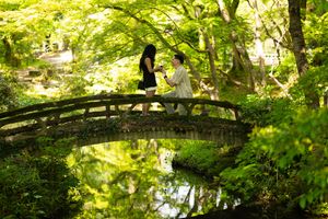 Couple stands on a rustic arched stone bridge in a lush green Japanese garden, with a pond reflecting the trees below.