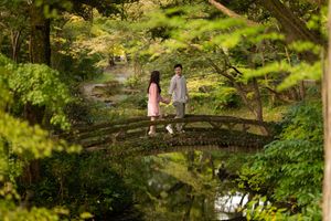 Couple in pink and gray holding hands while standing on a wooden arched bridge in a lush green Japanese garden.