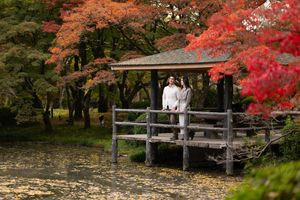 Couple standing on a wooden pavilion deck overlooking a calm pond, surrounded by vibrant red and orange autumn maple leaves in a Japanese ga