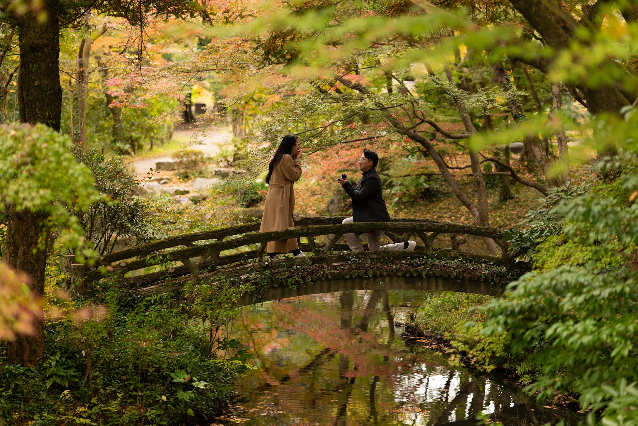 Couple standing on a curved wooden bridge over a calm pond in a lush Japanese garden surrounded by colorful autumn foliage.