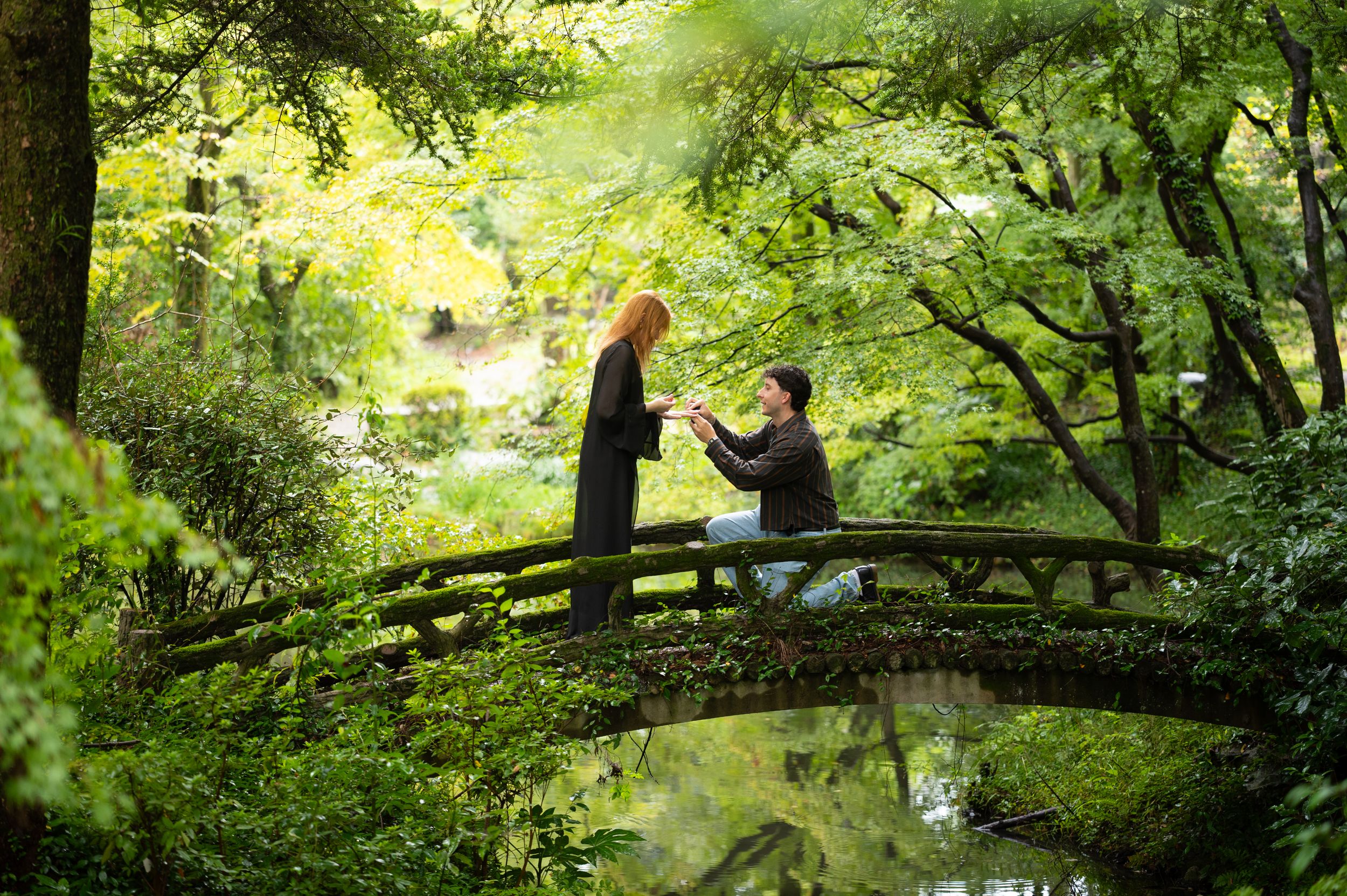 A man kneels on a wooden bridge proposing to a woman with red hair in a lush green Japanese garden.