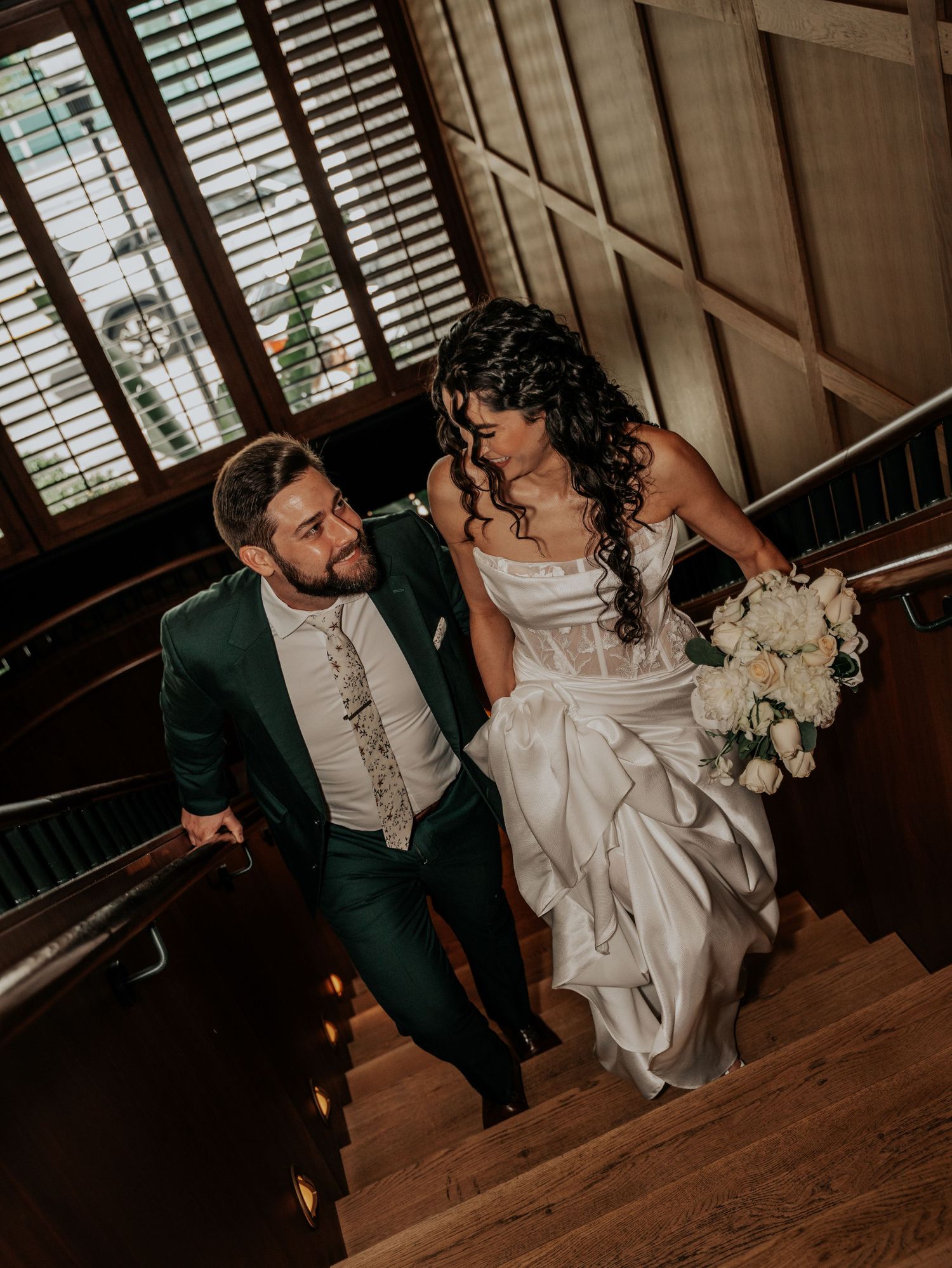 Bride in a white satin gown holding a white floral bouquet walks up a wooden staircase beside her groom in a green suit.
