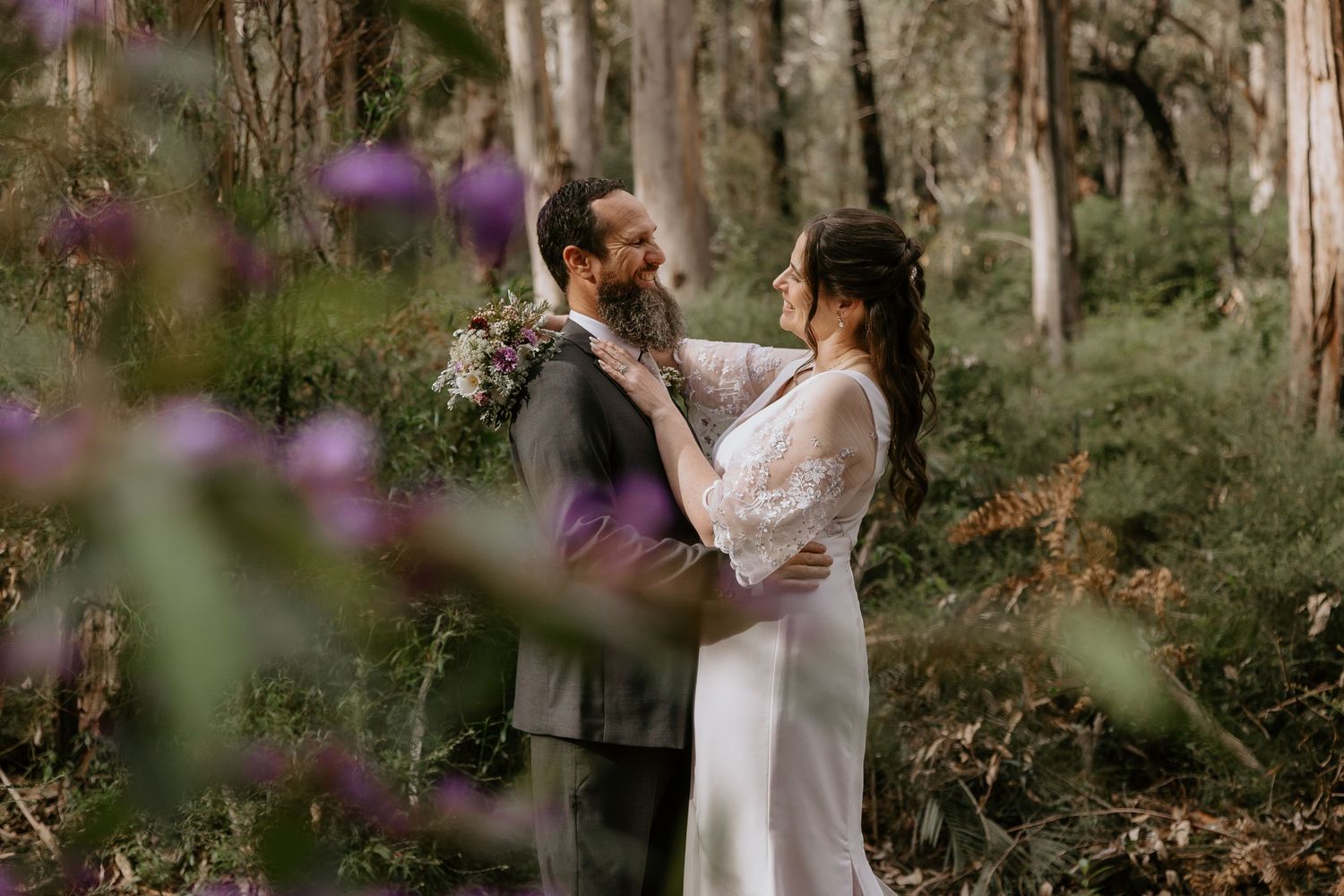 Bride in a white lace dress and bearded groom in a grey suit embrace in a sunlit bushland forest with purple wildflowers in the foreground.