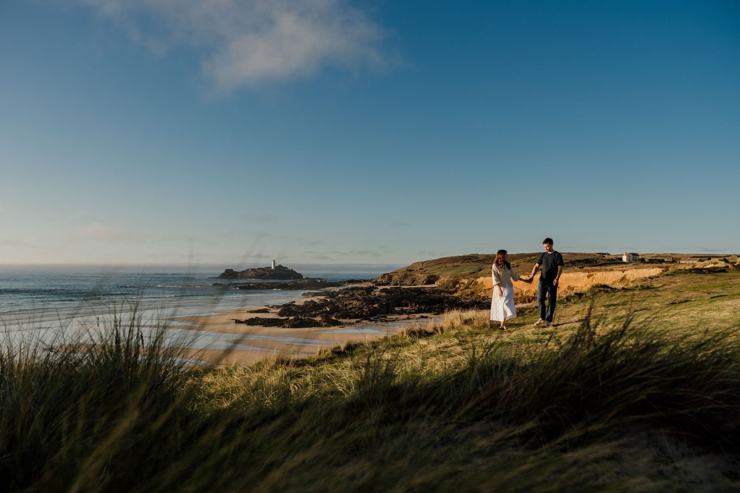Couple holding hands on coastal dunes with Godrevy lighthouse and beach in the background under a clear blue sky.