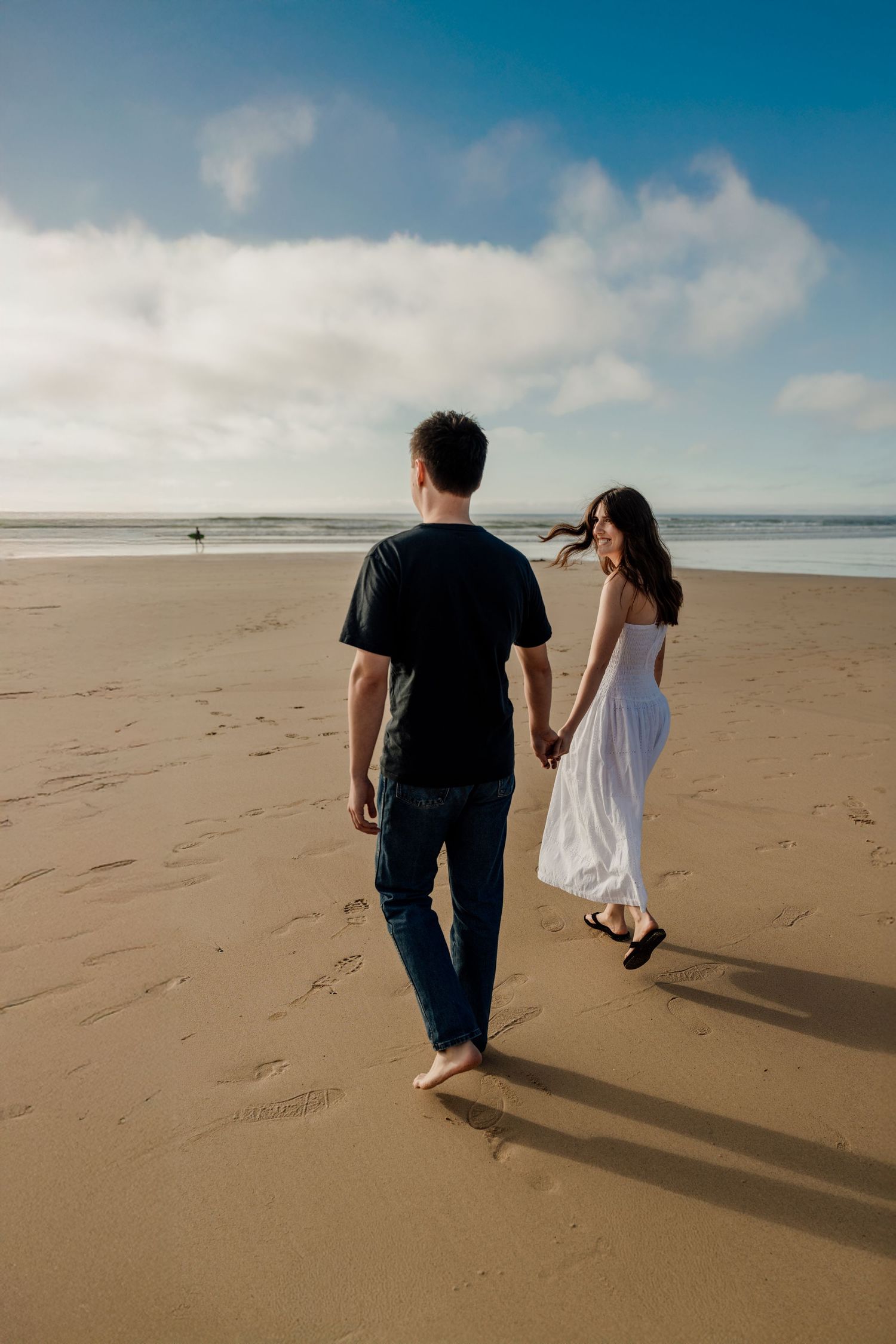 Couple holding hands walking on Godrevy beach, woman in white dress and man in black shirt, ocean and blue sky behind.