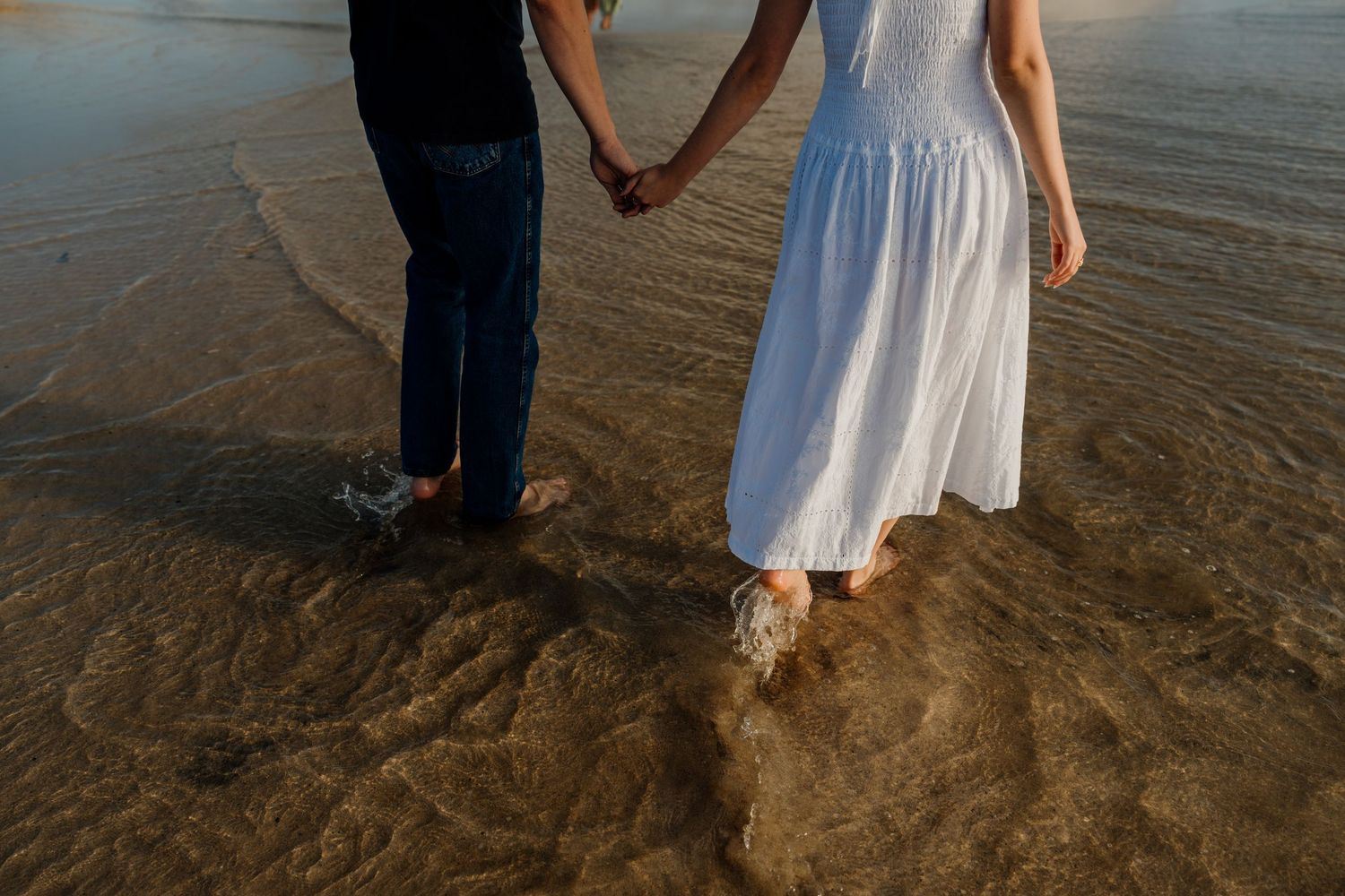 Couple holding hands barefoot on a sandy beach shore, woman in white dress and man in jeans.