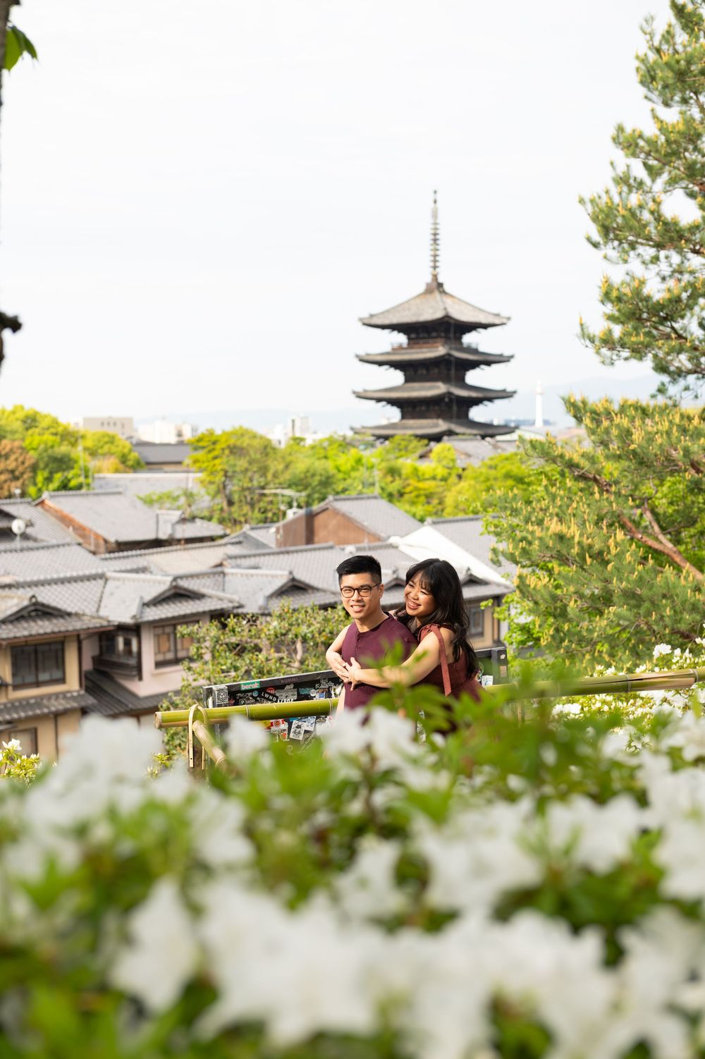 Couple embracing and smiling on a hillside overlooking traditional Kyoto rooftops and a five-story pagoda, framed by white azalea blooms.