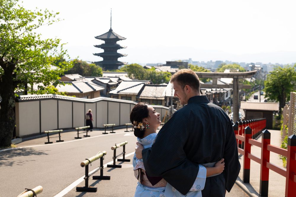 Couple embraces on a Kyoto hillside street, with a tall wooden pagoda, red railings and traditional rooftops in the background.