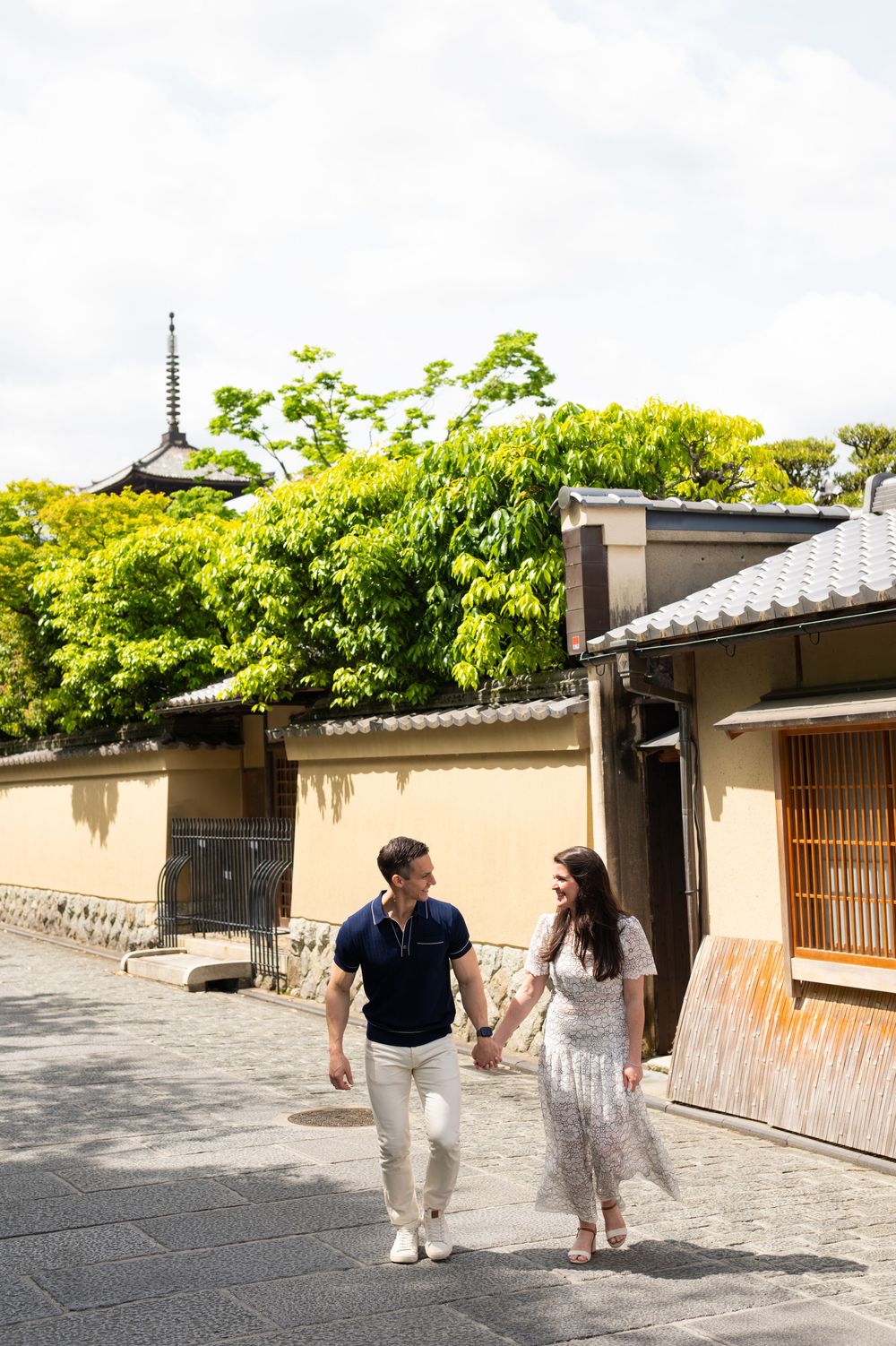 Couple holding hands and walking down a stone-paved Kyoto lane lined with traditional walls, with a pagoda rising above lush green trees.