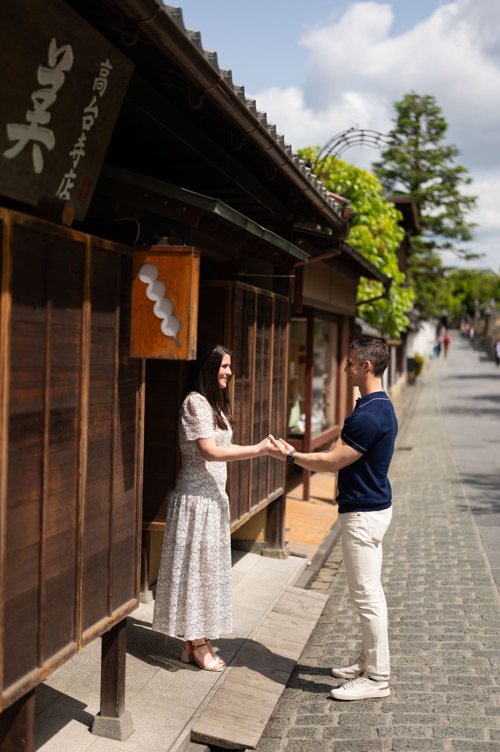 Couple holding hands beside a traditional wooden building with a hanging sign on a sunny cobblestone street in a historic Japanese town.