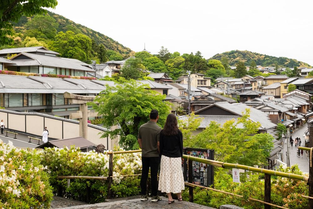 Couple standing at a wooden railing overlooking traditional Japanese rooftops and forested hills in a historic Kyoto neighborhood.