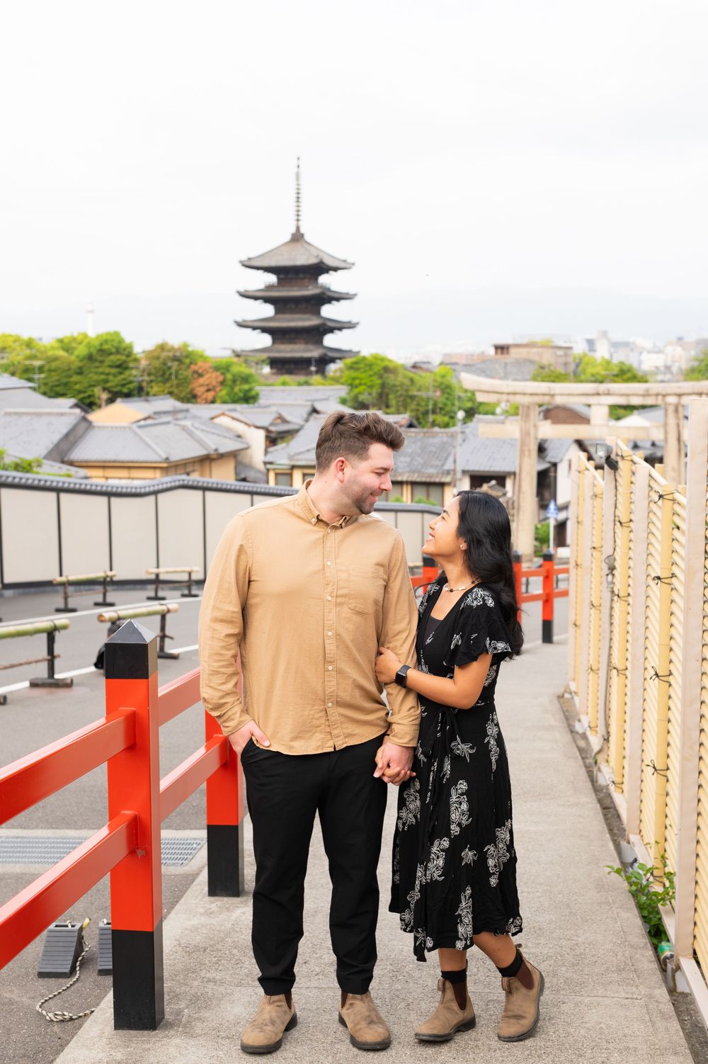 Smiling couple holding hands on a walkway with red railings and a historic Japanese pagoda in the background, Kyoto.