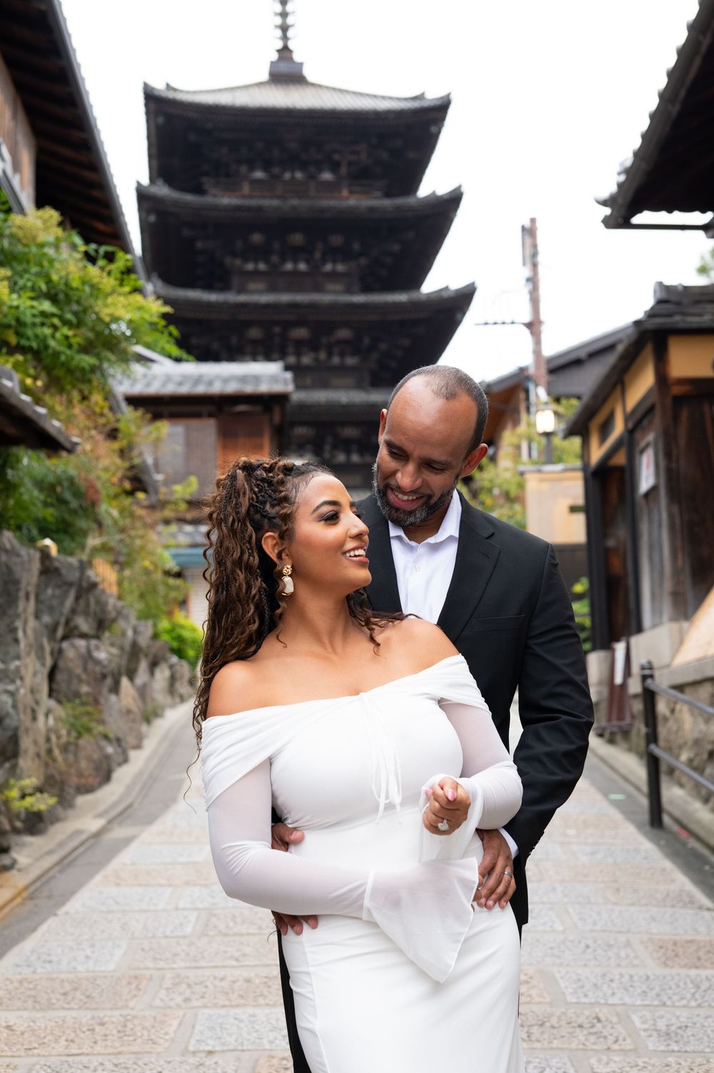 Couple in elegant white dress and dark suit embracing on a stone-paved lane with a historic Japanese pagoda in the background.