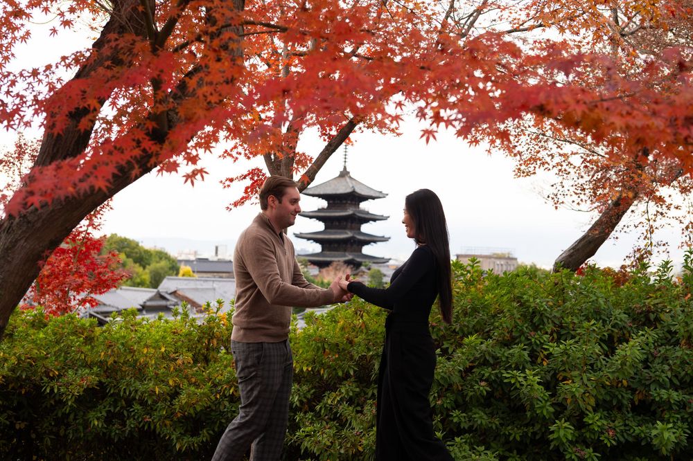 Couple holding hands under vibrant red autumn maple leaves with a Japanese pagoda and green hedges in the background.