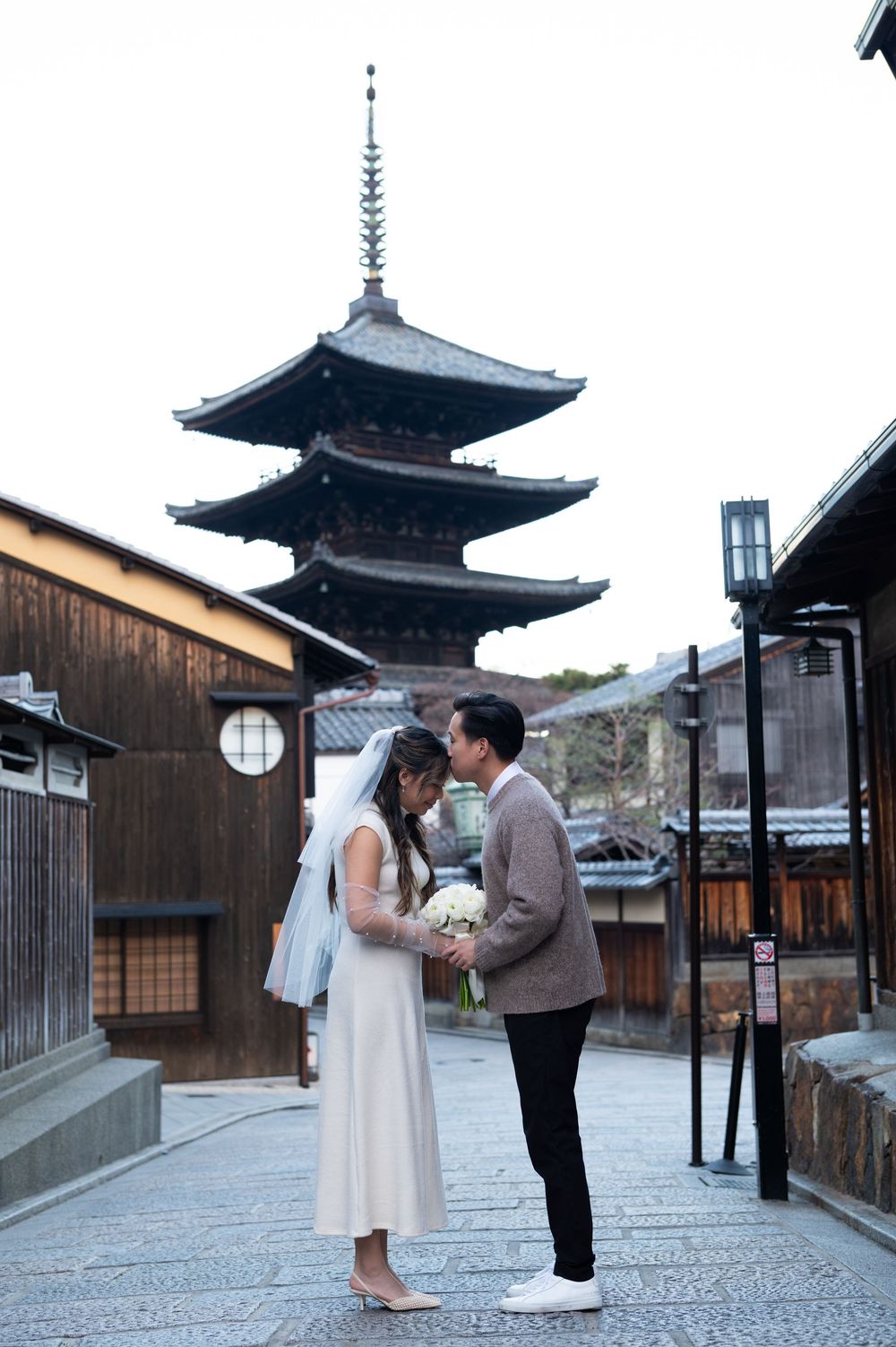 Bride in a white dress and veil holding a bouquet shares an intimate moment with her groom on a stone street near a historic Kyoto pagoda.
