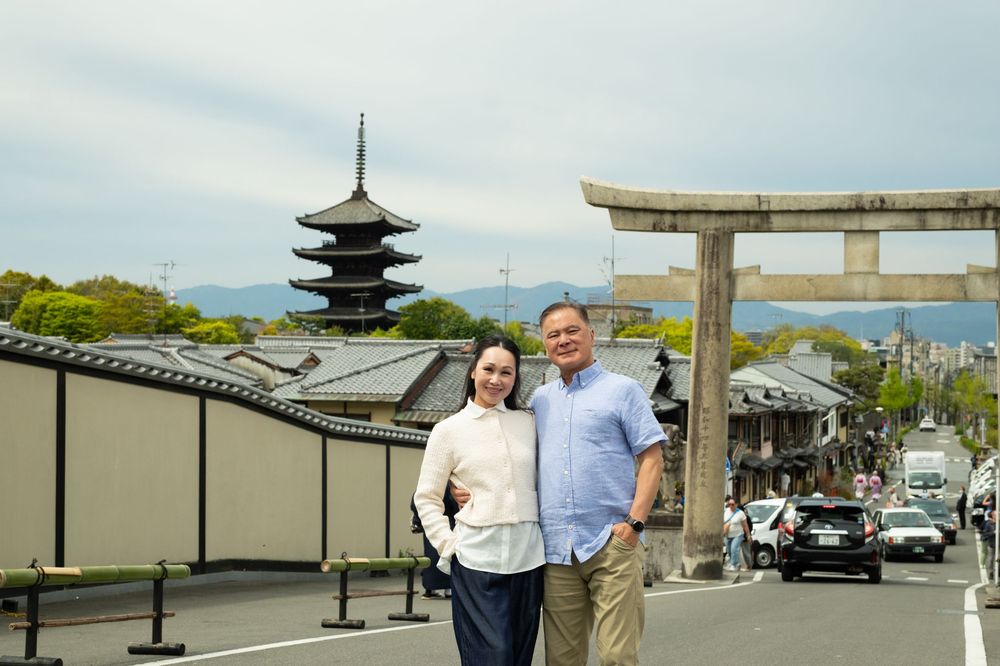 Smiling couple poses on a quiet Kyoto street with a traditional pagoda and stone torii gate behind them under a soft cloudy sky.
