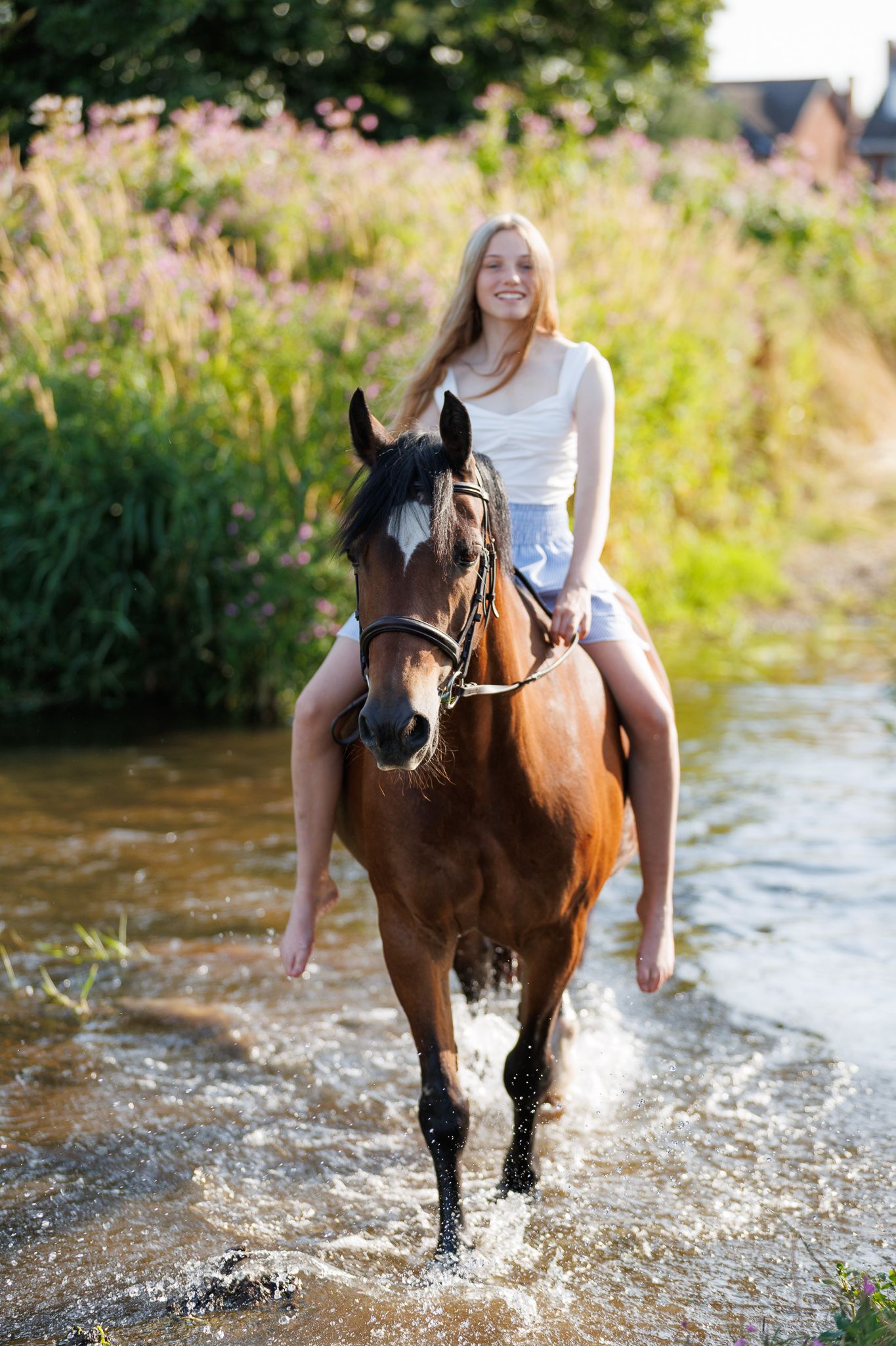 Smiling young woman with long hair rides a brown horse bareback through a shallow sunlit stream beside lush green summer vegetation.