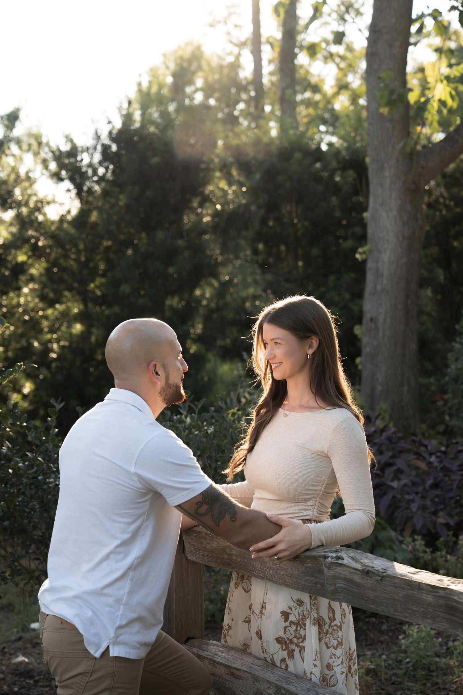 Engagement session photo of a couple smiling at each other beside a rustic wooden fence surrounded by lush green trees at sunset.