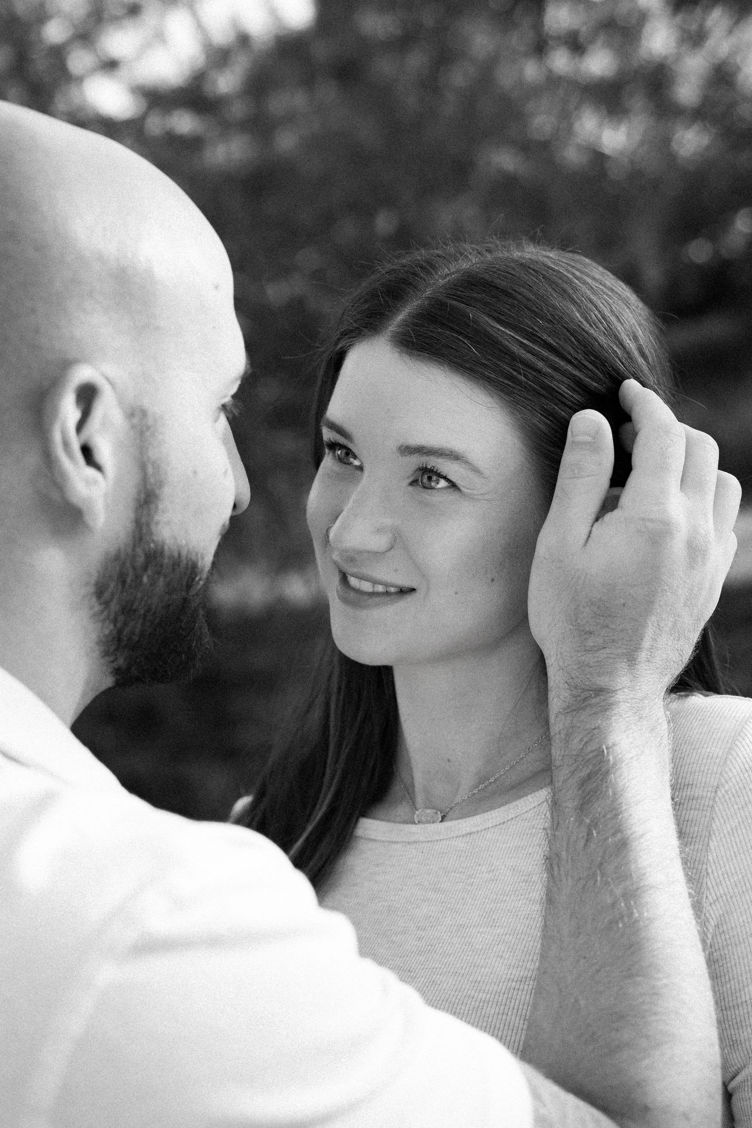 Black and white portrait of a smiling woman tucking her hair behind her ear while gazing at a bearded, bald man in a white shirt.