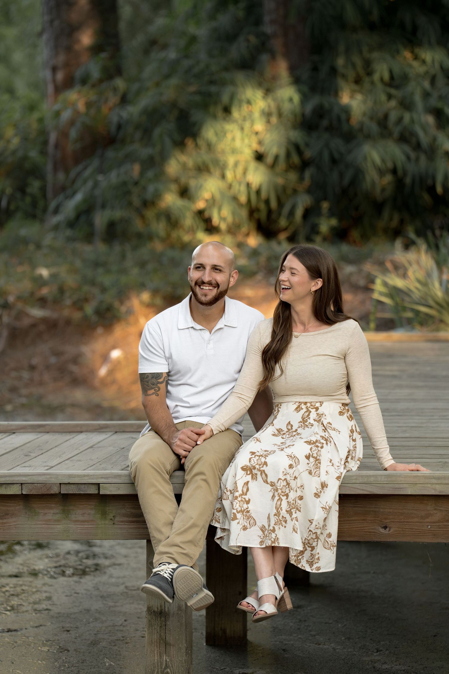 Smiling couple sitting together on a wooden dock surrounded by lush green trees, perfect for engagement or outdoor portrait photography.
