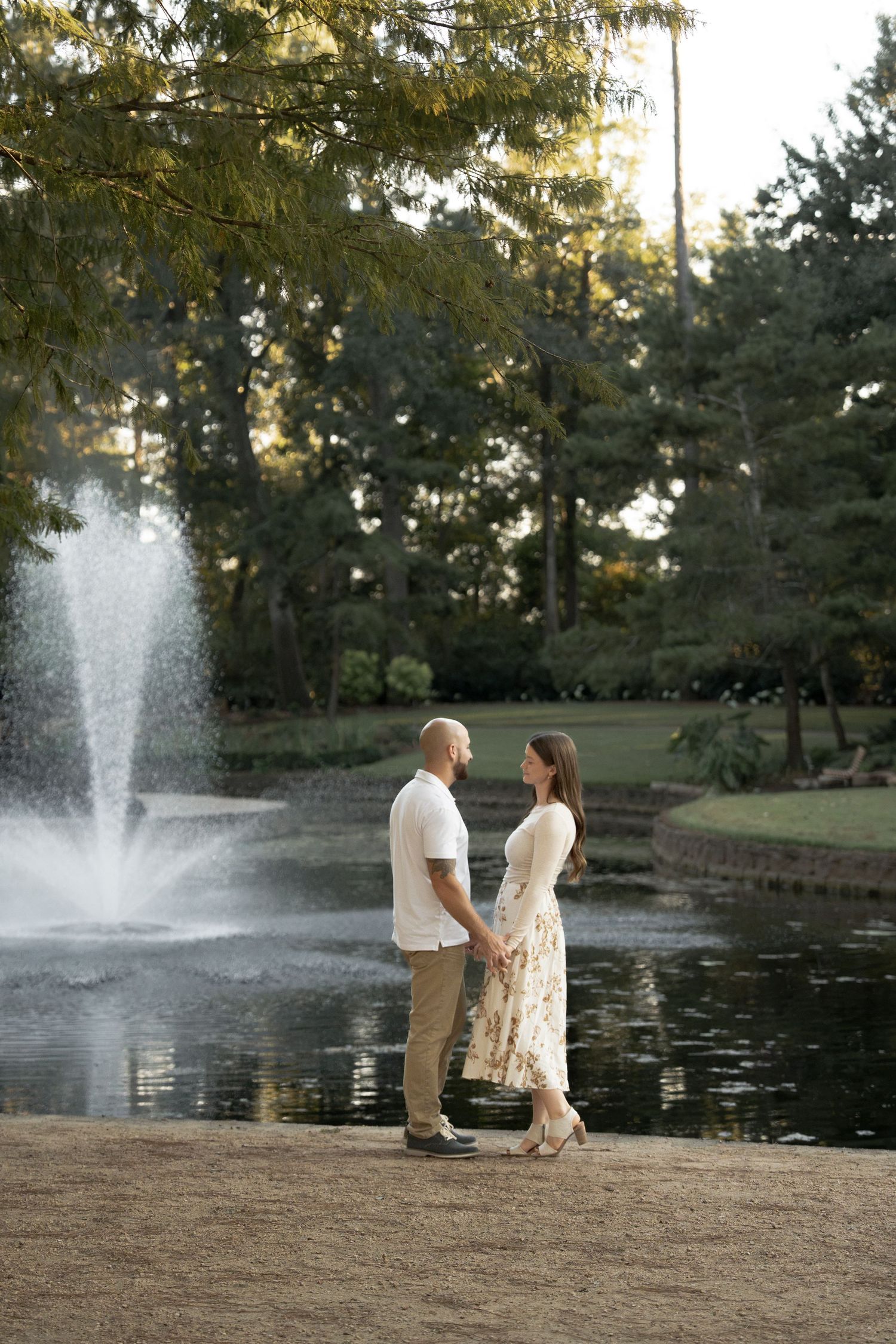 Couple holding hands and facing each other beside a park pond with a fountain, surrounded by tall trees in warm evening light.